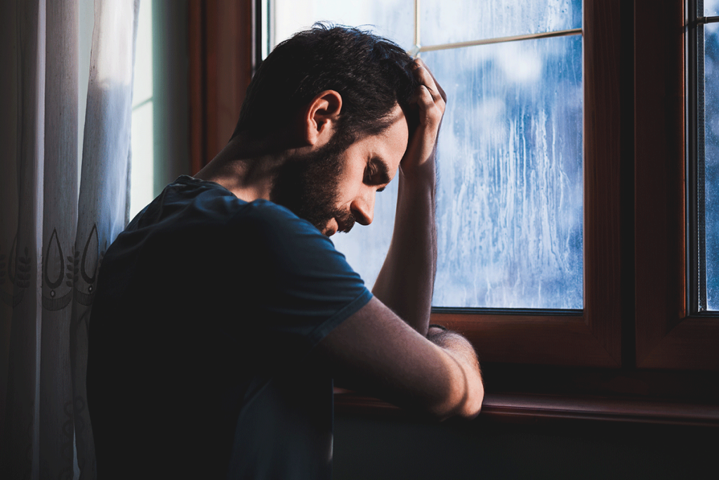 a person living with signs of depression holds their head as they look out a window sadly